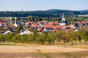 Das Bild zeigt die Orstansicht der Gemeinde Wehrheim im Taunus mit der katholischen und der evangelischen Kirche. Im Vordergrund ist ein Weizenfeld zu sehen, dahinter Streuobstbäume.