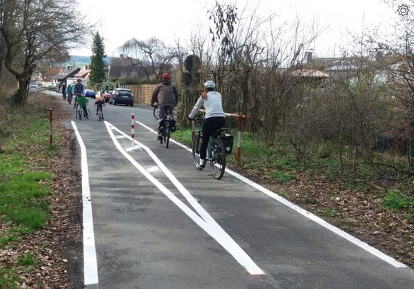 Das Foto zeigt einen Radfahrweg mit aufgemalten Markierungen, auf dem Radfahrende in beide Richtungen fahren, in Wehrheim.