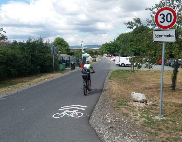Radfahrer auf Straße am Schwimmbad mit Radpiktogramm